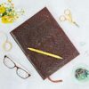 The Large Brown Embossed Notebook is decorated with a yellow pen and surrounded by glasses, scissors, a bowl of paperclips, and a small bouquet on a light surface.
