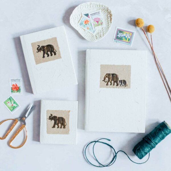 Three Elephant Dung Notebooks, adorned with charming illustrations, are set on a white surface amid scissors, twine, a plate holding stamps, and two yellow flowers.