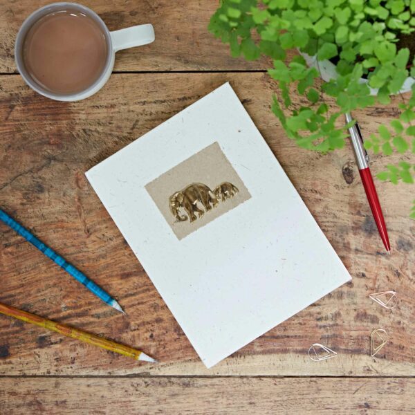 Wooden table with a cup of tea, a small plant, paperclips, colored pencils, and a notebook featuring gold elephants on the cover.
