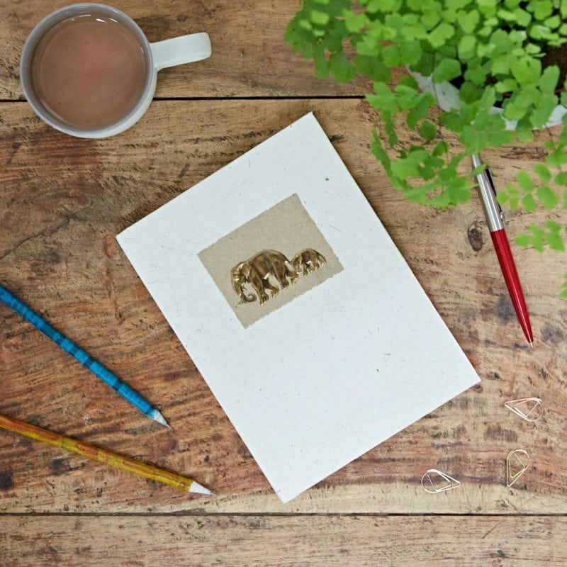 Wooden table with a cup of tea, a small plant, paperclips, colored pencils, and a notebook featuring gold elephants on the cover.