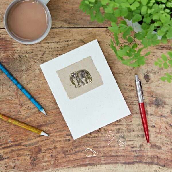 A notebook with an elephant design is on a wooden table, surrounded by a cup of coffee, two pencils, a pen, three paper clips, and a green plant.