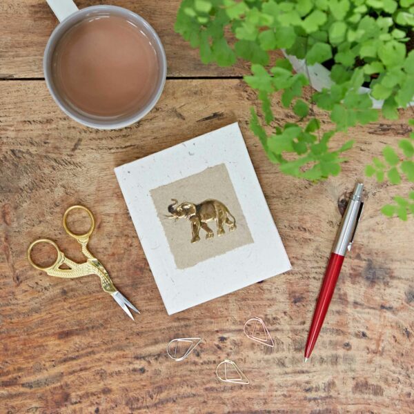 A cup of coffee, gold scissors, three paperclips, a red pen, and a handmade card with an elephant design are on a wooden table next to a green plant.