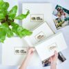 A person holds the Elephant Dung Photo Album featuring a gold elephant design, surrounded by similar albums, a green plant, and printed photos.