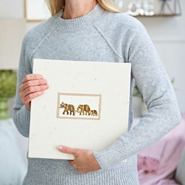 A person in a gray sweater holds the Elephant Dung Photo Album, which features a white cover adorned with a gold illustration of four elephants.