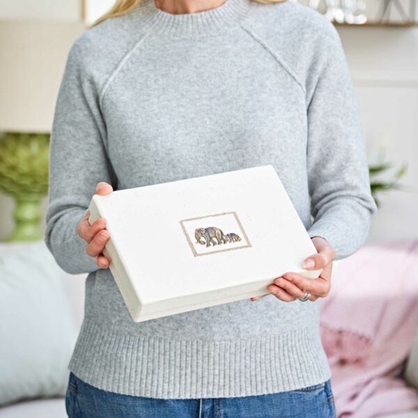 A person in a gray sweater and blue jeans holds the Elephant Dung Photo Album—a white box with an elephant illustration on the lid.