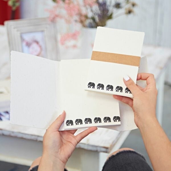 Hands holding an Elephant Dung Letter Writing Set with an elephant design, framed by a photo and flowers in the background.