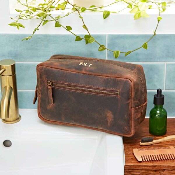 A brown leather wash bag with initials "P.R.T." sits by a sink, next to a green bottle, comb, and brush, with plants above and blue tiled wall in the background.
