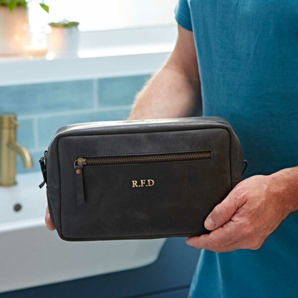 A person in a blue shirt holds a Personalised Buffalo Leather Wash Bag—an eco-friendly black leather toiletry bag with gold "R.F.D." initials above the front zip pocket—while standing in a bathroom.