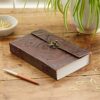 An Indra Extra Large Embossed Leather Journal with a clasp sits on a wooden table. Nearby are a pen, a bowl of paper clips, and a potted plant.