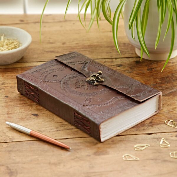 An Indra Extra Large Embossed Leather Journal with a clasp sits on a wooden table. Nearby are a pen, a bowl of paper clips, and a potted plant.