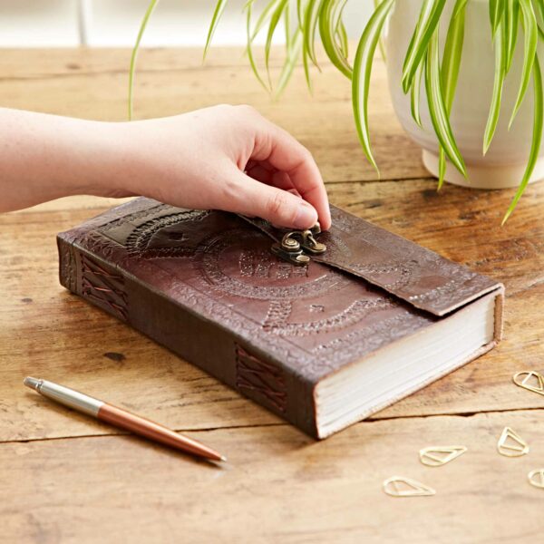 A hand is unfastening a metal clasp on a closed, Indra Extra Large Embossed Leather Journal on a wooden surface. Nearby are a potted plant, a pen, and gold paperclips.