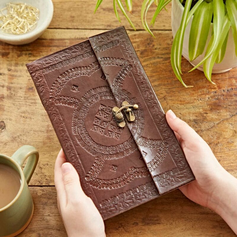 Hands holding an Indra Extra Large Embossed Leather Journal with a brass clasp, placed on a wooden table alongside a bowl of paperclips, a green plant, and a cup of tea.