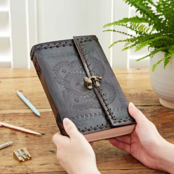Hands holding a closed Indra Extra Large Embossed & Stitched Leather Journal with a clasp, placed on a wooden table next to pens, a pencil sharpener, and a potted fern.