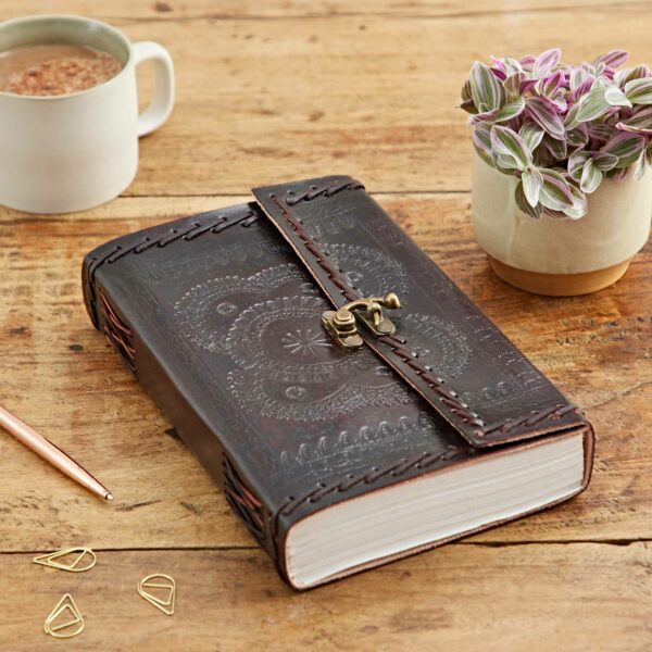 An Indra Extra Large Embossed & Stitched Leather Journal with a dark brown leather cover and metal clasp lies on a wooden table. Nearby are a pen, paperclips, a mug, and a small potted plant.