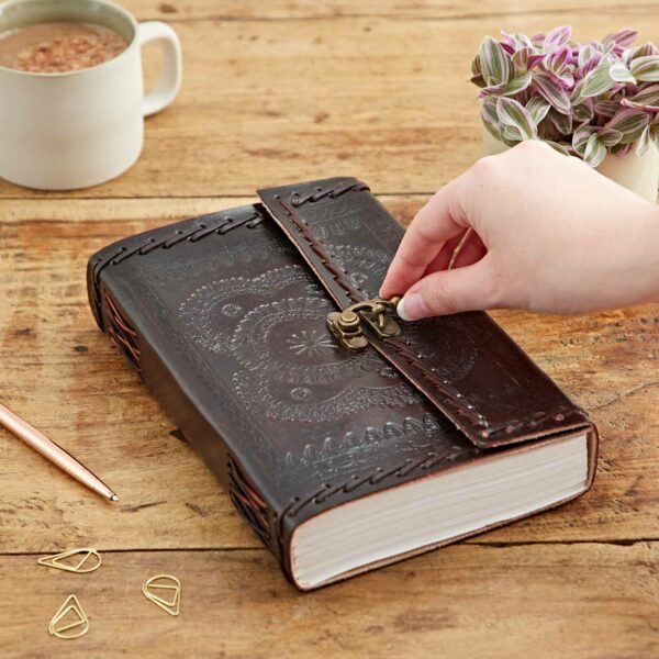 A hand opening the clasp of an Indra Extra Large Embossed & Stitched Leather Journal on a wooden table, with a pen, paper clips, a potted plant, and a mug in the background.