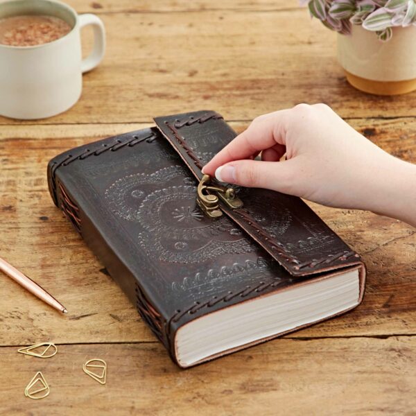 An individual opening the Indra Extra Large Embossed & Stitched Leather Journal with a brass clasp on a wooden table, next to a mug, a pen, and some paper clips.
