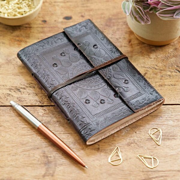 An Indra Embossed Leather Notebook with a wraparound strap is on a wooden table next to a silver and rose gold pen, a dish of pasta, three gold paperclips, and a small potted plant.
