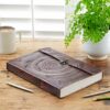 An Indra A4 Embossed Leather Journal rests on a wooden table next to a potted plant. A mug and two pens are also on the table, completing the serene setting for reflection.