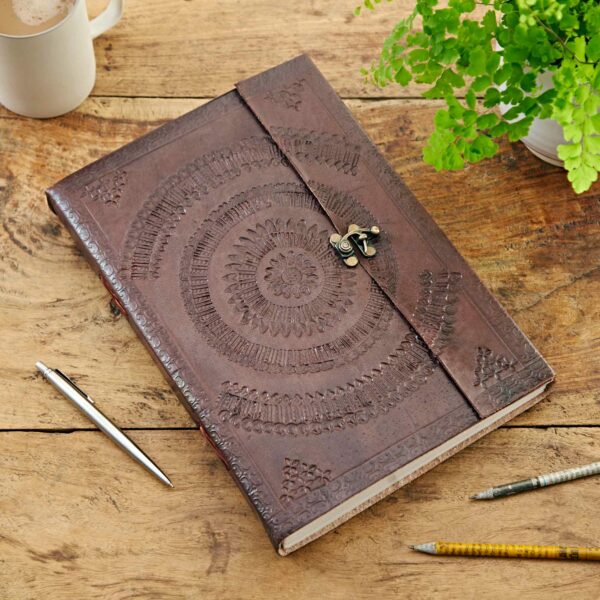 A closed Indra A4 Embossed Leather Journal with an ornate embossed cover and clasp sits on a wooden table next to pens, a pencil, a plant, and a coffee mug.