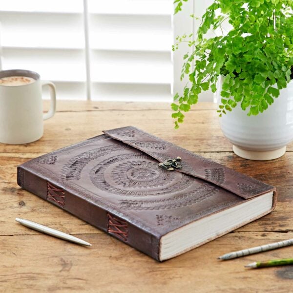 An Indra A4 Embossed Leather Journal rests on a wooden table next to a potted plant. A mug and two pens are also on the table, completing the serene setting for reflection.