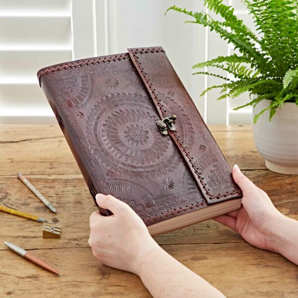 A person holding a large, embossed and stitched leather journal with a clasp, beside pencils, a sharpener, and a potted fern on a wooden table. The elegance of the Indra A4 Embossed & Stitched Leather Journal shines through its meticulous craftsmanship.