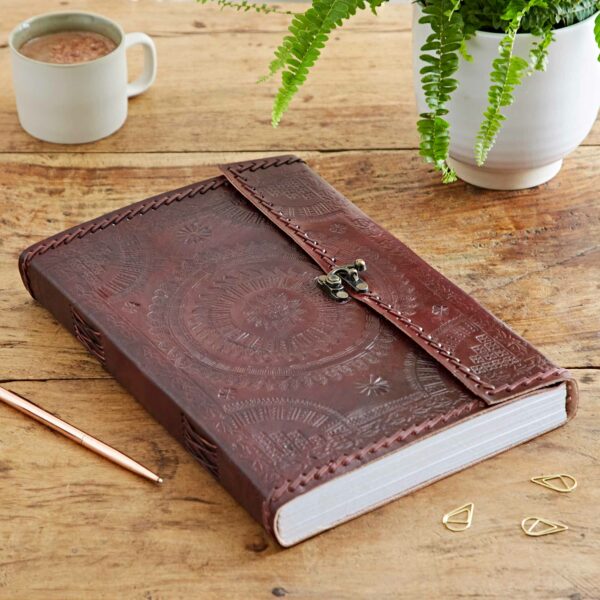 The Indra A4 Embossed & Stitched Leather Journal, featuring a secure clasp, lies closed on a wooden table alongside a cup of coffee, a pen, several paper clips, and a potted fern.