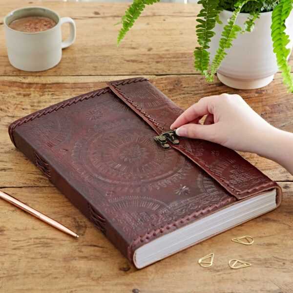 A hand opens the clasp on an Indra A4 Embossed & Stitched Leather Journal resting on a wooden table. Nearby are a cup of coffee, a fern plant, a pen, and paper clips.