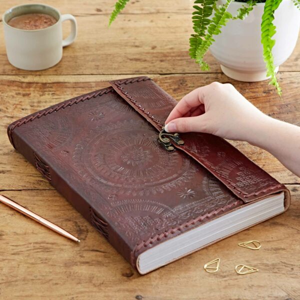 A hand is opening a clasp on an Indra A4 Embossed & Stitched Leather Journal on a wooden table. Nearby are a cup of coffee, a plant, a pen, and some paper clips.