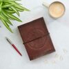 A Celtic Star Leather Journal with a pentagram design, a red pen, a coffee mug, a plant, and paperclips are arranged on a white surface.