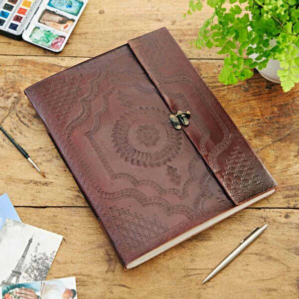 A large, decorative Indra Extra Large Embossed Leather Photo Album lies closed on a wooden table. Nearby are watercolor paints, a pen, a small green plant, and a photo of the Eiffel Tower.