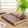 An Indra Extra Large Embossed Leather Photo Album with a clasp sits on a wooden table beside a pot of green fern, photographs, a pen, and a watercolor paint set.
