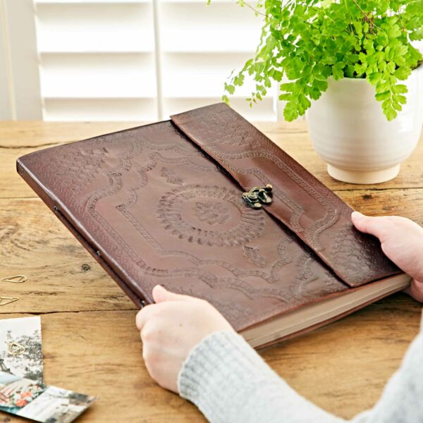 A person holds an Indra Extra Large Embossed Leather Photo Album on a wooden table adorned with a potted plant and scattered photographs. The embossed leather cover adds an elegant touch, making it resemble a cherished photo album.