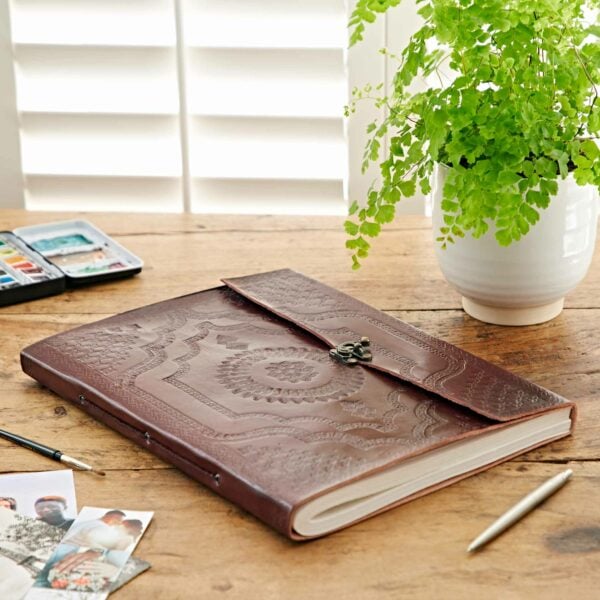 An Indra Extra Large Embossed Leather Photo Album with a clasp sits on a wooden table beside a pot of green fern, photographs, a pen, and a watercolor paint set.