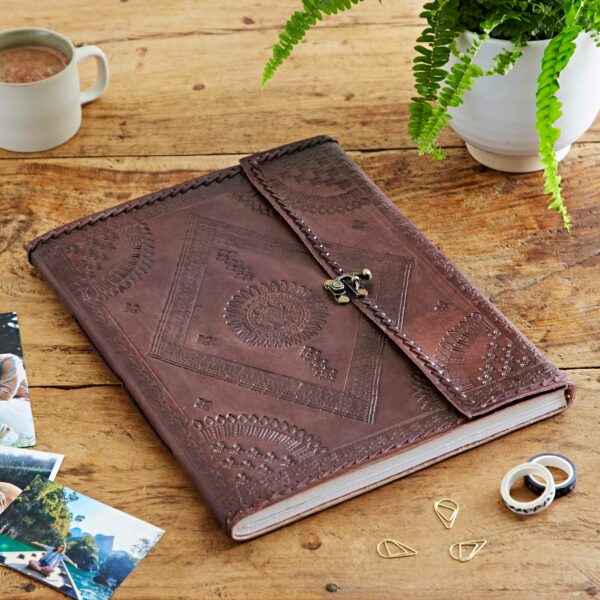 The Indra Extra Large Embossed & Stitched Leather Photo Album with an ornate clasp lies on a wooden table, surrounded by a plant, a mug, scattered photos, and a few decorative rings and paper clips. Its embossed leather cover gives it the look of a cherished journal.