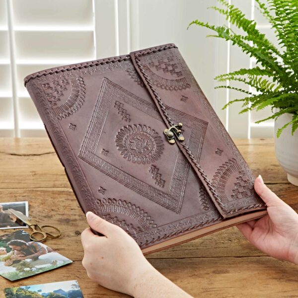 A person holds an Indra Extra Large Embossed & Stitched Leather Photo Album with a clasp. Nearby, there are photos and a pair of scissors on a wooden table. A potted fern is in the background.