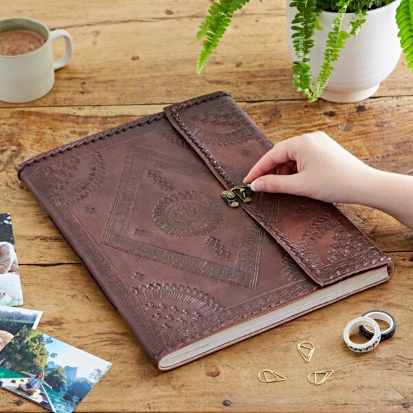 A hand opens an Indra Extra Large Embossed & Stitched Leather Photo Album on a wooden table. Nearby are photos, a mug, plant, paper clips, and rolls of washi tape.