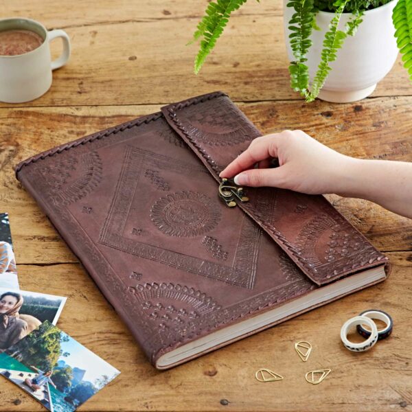 A hand is opening a clasp on an intricately designed Indra Extra Large Embossed & Stitched Leather Photo Album. The journal is on a wooden table with photos, paper clips, a mug, and a potted fern nearby.
