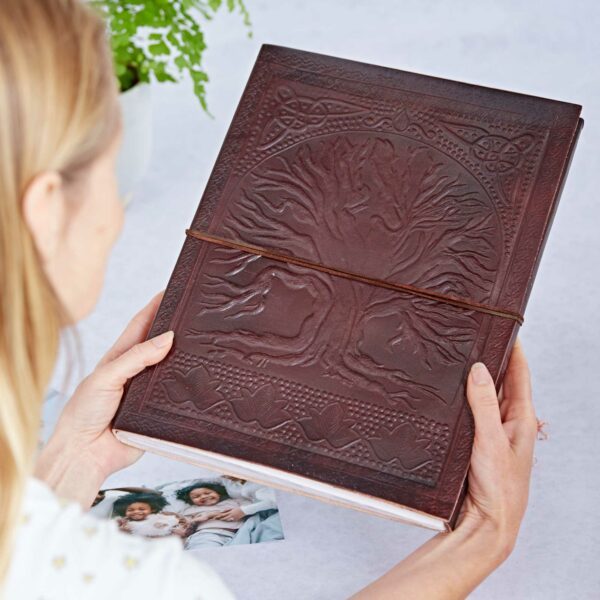 A person holds a large leather-bound journal, its cover graced with an intricate Tree of Life embossing. Nearby, two photos rest on the table, waiting to find their place in the elegant leather photo album.