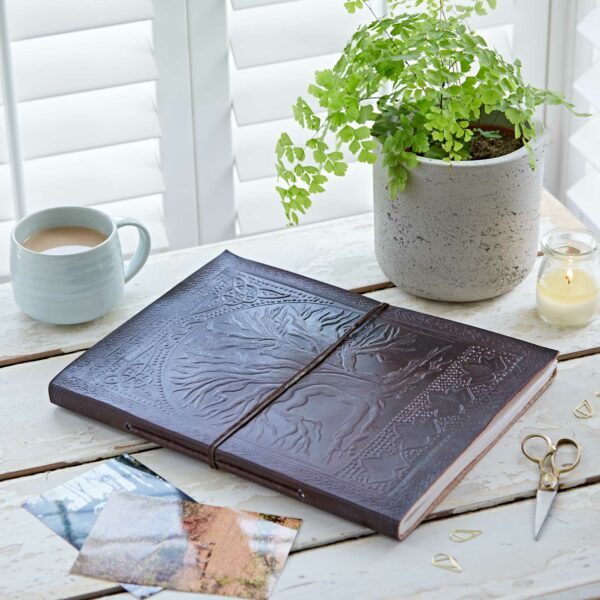 A large Leather Tree of Life Embossed Photo Album sits on a wooden table with a potted plant, tea, candle, photos, scissors, and small gold paperclips.