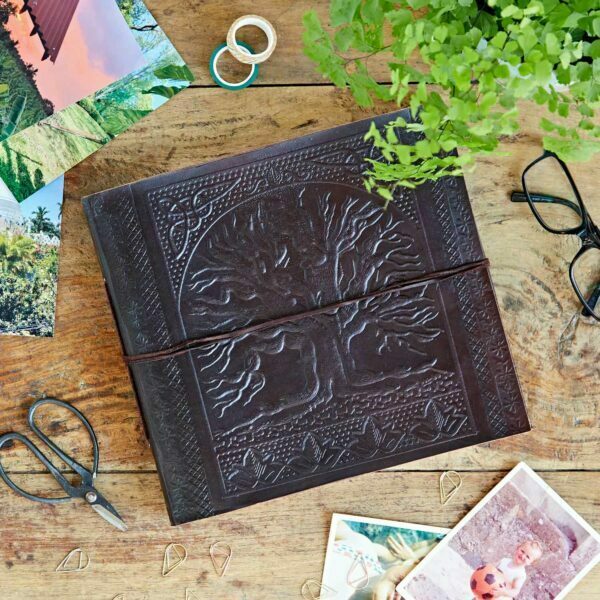 A Leather Tree of Life Embossed Photo Album sits on a wooden table, surrounded by photos, glasses, scissors, paperclips, rings, and a plant.