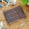 A leather-bound journal with an embossed cover, placed on a wooden table surrounded by craft supplies including photos, scissors, tape, and paper clips. A small potted plant is also visible. The scene could easily be mistaken for a cozy moment of making an Indra Medium Embossed Leather Photo Album.