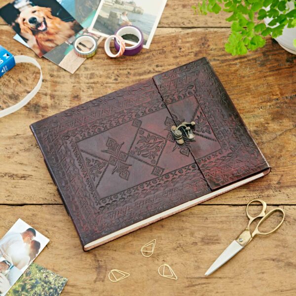 A leather-bound journal with an embossed cover, placed on a wooden table surrounded by craft supplies including photos, scissors, tape, and paper clips. A small potted plant is also visible. The scene could easily be mistaken for a cozy moment of making an Indra Medium Embossed Leather Photo Album.