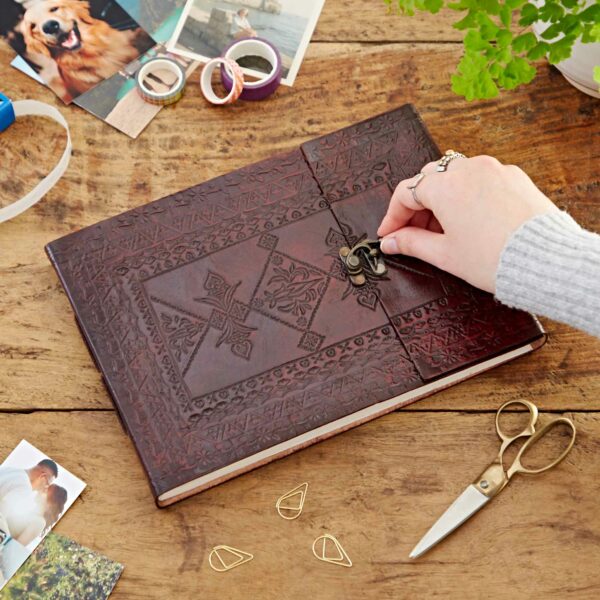 A hand unfastens a decorative latch on an Indra Medium Embossed Leather Photo Album placed on a wooden table, surrounded by photos, scissors, tape, paperclips, and a potted plant.