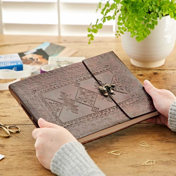 A person holds an intricately decorated brown leather journal with a clasp on a wooden table, evoking the timeless charm of an Indra Medium Embossed Leather Photo Album. Nearby, paper clips, scissors, a plant in a white pot, and scattered photos and stationery items enhance the creative ambiance.