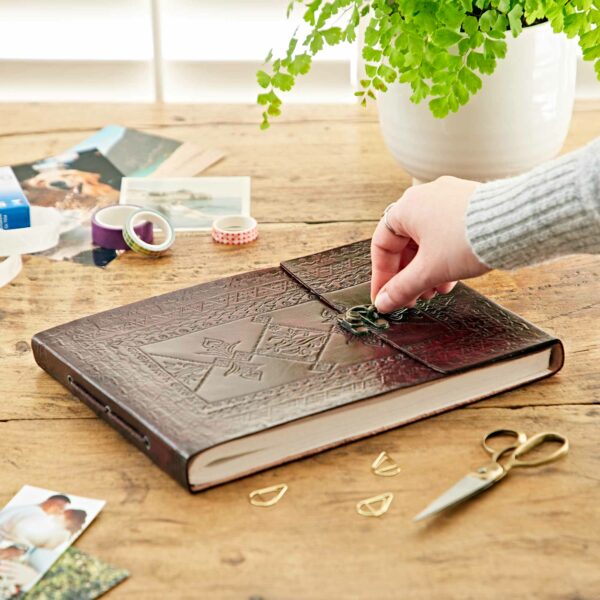 A hand reaches to open a clasped, Indra Medium Embossed Leather Photo Album on a wooden table surrounded by photos, washi tape, a potted fern, and crafting supplies.