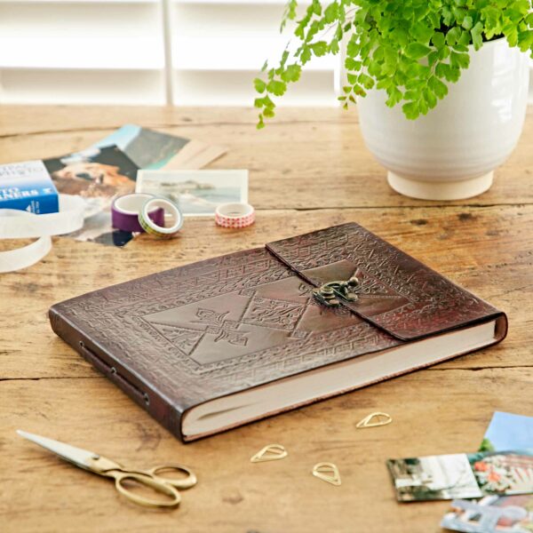 An Indra Medium Embossed Leather Photo Album rests on a wooden table along with photographs, scissors, tape, paper clips, and a potted plant in the background.