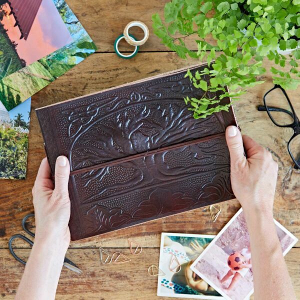 Hands gently cradle an M Size leather photo album embossed with the Tree of Life. Surrounding it on the wooden table are glasses, a plant, scissors, photos, paperclips, and a roll of tape—each item adding to the cozy atmosphere of creative nostalgia.