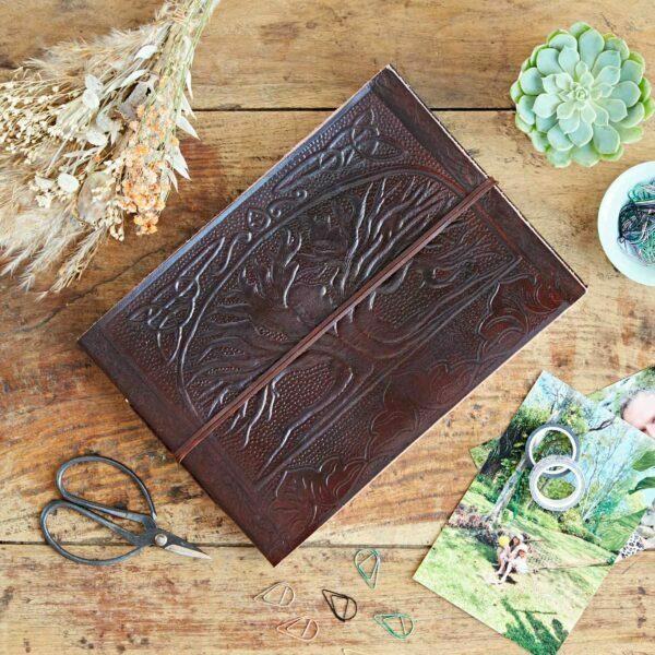A Leather Tree of Life Embossed Photo Album rests on a wooden table, surrounded by dried flowers, a succulent, scissors, colorful paperclips, and photographs.