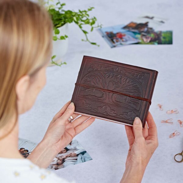 A person holding a brown leather journal with a Tree of Life design, surrounded by scattered photos, paperclips, and a small plant on a light surface.