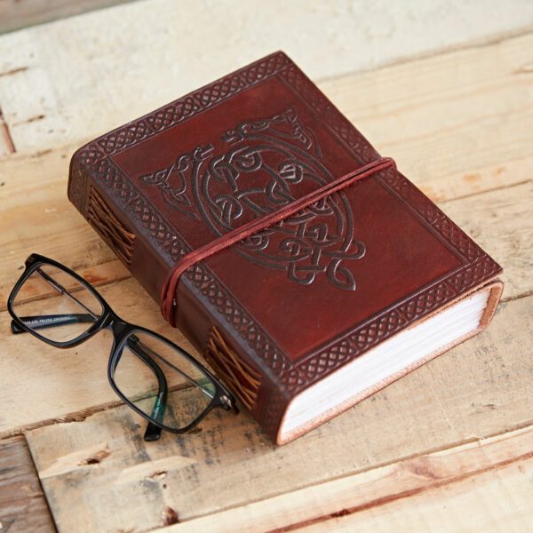 The Celtic Serpent Leather Journal, a closed embossed leather notebook with a cord, lies on a wooden surface next to black eyeglasses.